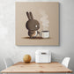 Grand tableau sur le mur d'une cuisine au style japonaise et minimaliste. Un lapin marron malicieux, le regard figé par la douleur a été pris au dépourvu par la chaleur de la tasse de café qu'il tente de saisir. Aux couleurs taupe et beige, la scène humoristique apporte à cela mouvement et vitalité