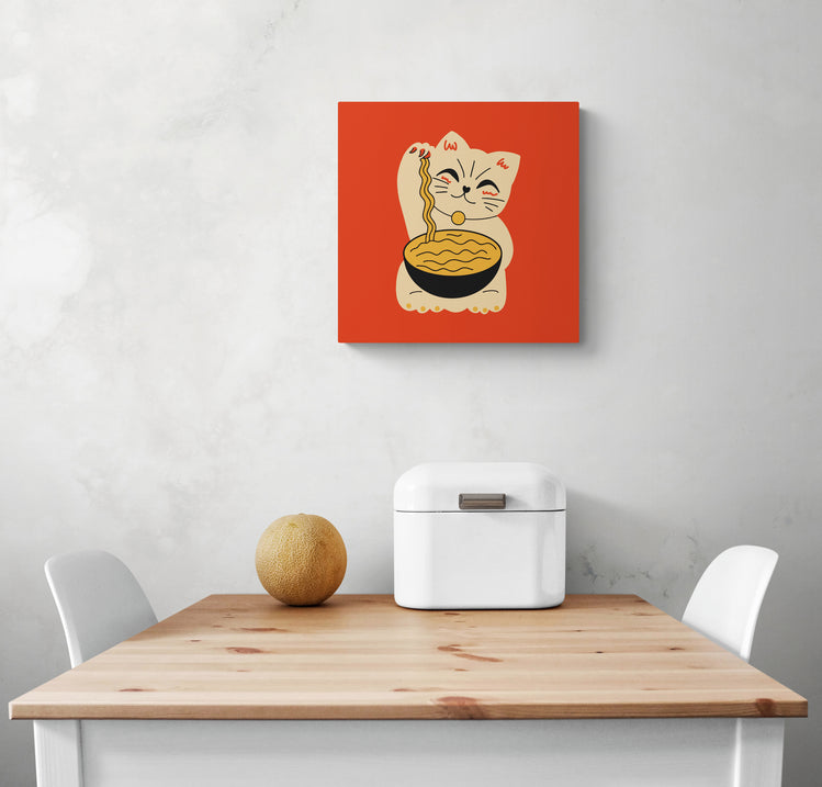 Un petit tableau cuisine avec un chat maneki-neko accroché sur un mur blanc. Dessous une table de repas et deux chaises blanches. Un melon et une boîte à pain sont sur la table.