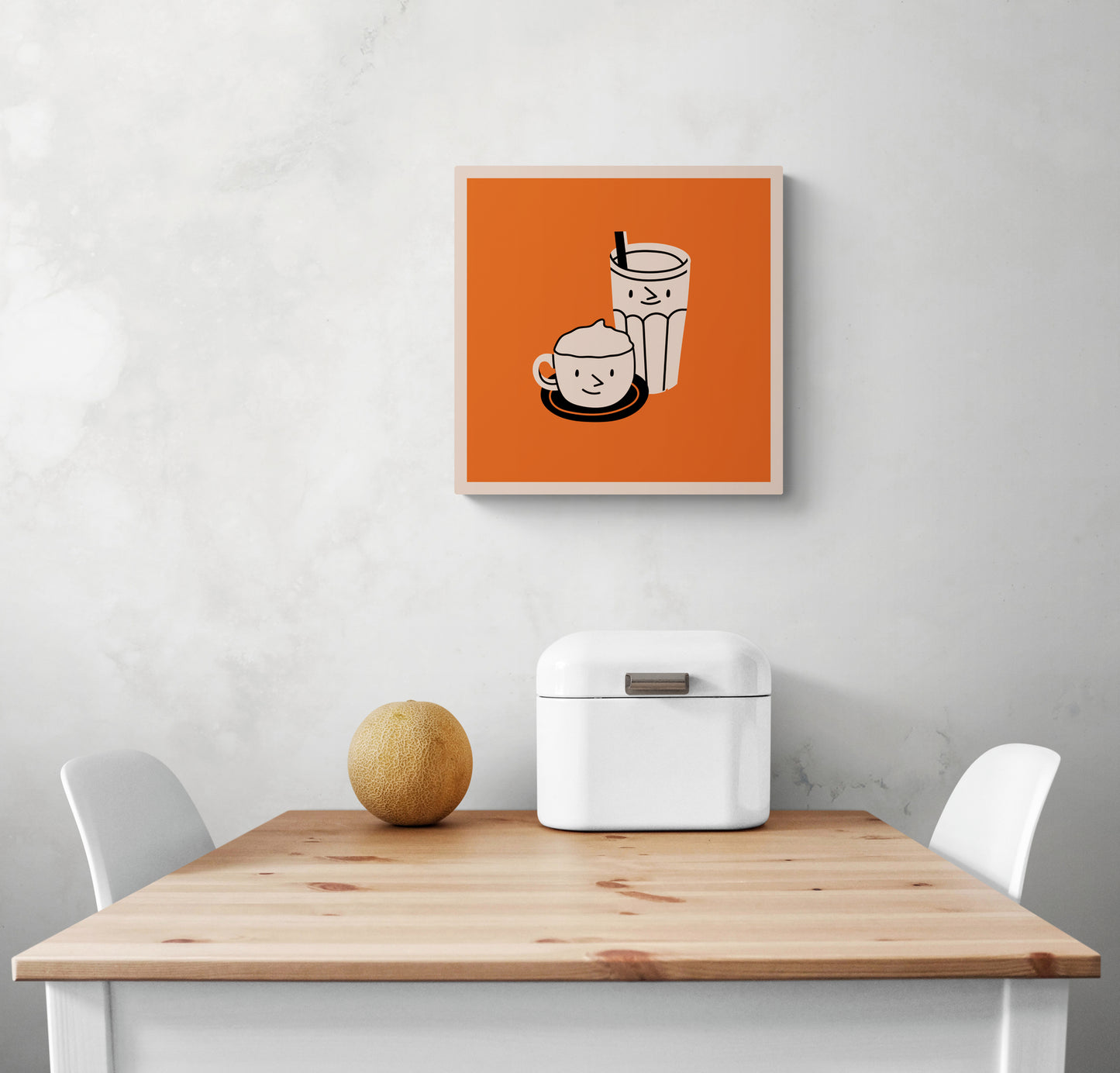  Un Tableau décoration murale orange dans une cuisine blanche. Ce tableau cuisine est placé au-dessus d'une table en bois et deux chaises sont de chaque côté. Une boîte à pain en métal blanc et un melon sont sur la table
