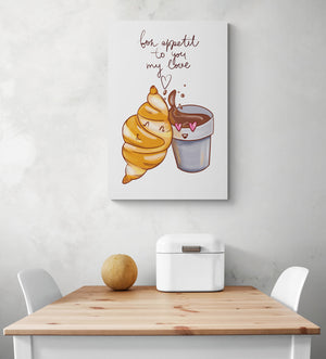 Un tableau décoration cuisine avec une l'illustration d'un croissant et chocolat chaud est accroché sur le mur blanc. Cette toile est placée au-dessus d'une table en bois et deux chaises sont de chaque côté. Une boîte à pain en métal blanc et un melon sont sur la table