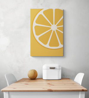 grand tableau coloré orange habille un mur blanc. Le tableau represente une illustration d'une tranche d'orange. En dessous du tableau, on peut apercevoir une table en bois et deux chaises blanches qui semblent être placées là pour offrir un espace confortable. Sur la table, il y a une corbeille à pain en métal blanc et un petit melon vert.