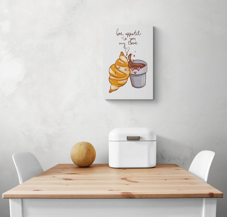 Un petit tableau blanc avec une illustration enfantine est accroché au mur d'une cuisine blanche. Ce tableau cuisine est placé au-dessus d'une table en bois et deux chaises sont de chaque côté. Une boîte à pain en métal blanc et un melon sont sur la table