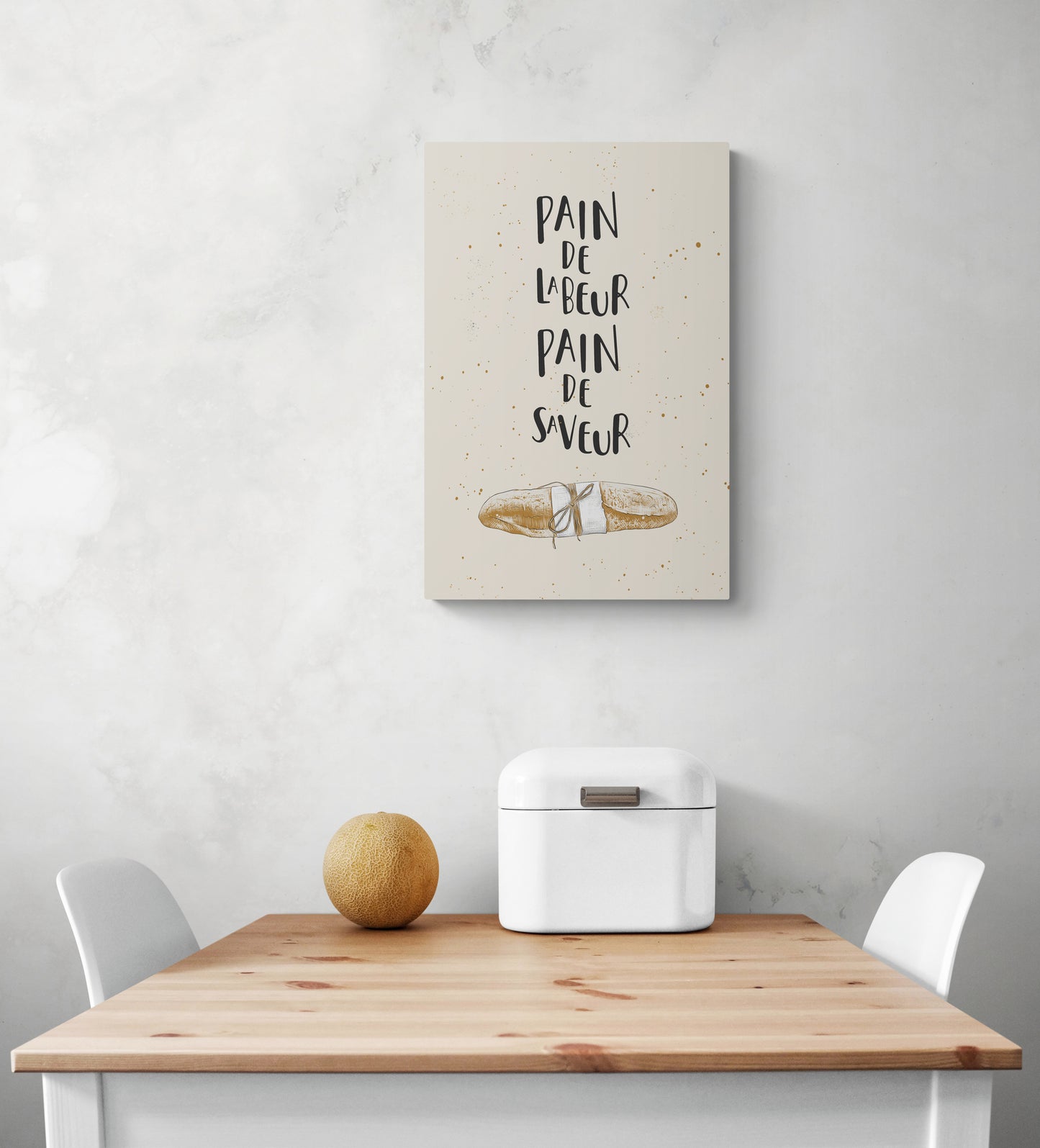 Un tableau mural cuisine beige habille un mur blanc. Sur ce tableau une baguette et une citation. En dessous du tableau, une table en bois et deux chaises blanches à chaque extrémité