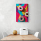 Un tableau décoration cuisine coloré avec trois donuts est accroché sur le mur blanc. Ce tableau cuisine est placé au-dessus d'une table en bois et deux chaises sont de chaque côté. Une boîte à pain en métal blanc et un melon sont sur la table