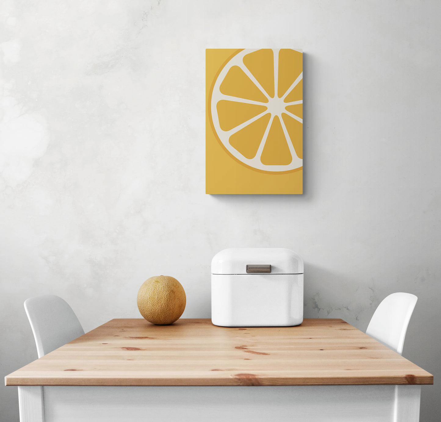  un petit tableau décoratif mural est accroché sur le mur blanc d'une cuisine. En dessous se trouvent une table en bois doré et deux chaises blanches. Sur la table se trouvent une corbeille à pain et un petit melon. Tous ces éléments créent un look coloré et harmonieux dans la pièce.