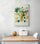A decorative kitchen art piece hanging on a white wall. It features a cluster of green grapes surrounded by green leaves and lemon slices. This art brings a fresh and gentle color palette. Below, a pine dining table with two white chairs facing each other.