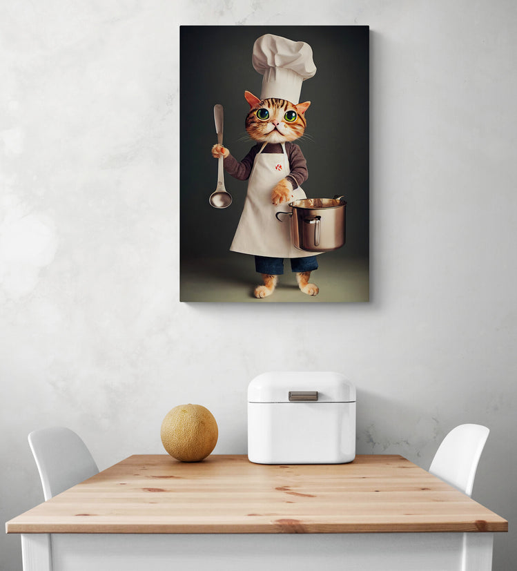 Dans une cuisine, sur un mur blanc avec des nuance de gris il y a un tableau qui represente un chat de plein pieds vêtu d’un uniforme et d’une toque de cuisinier dans ces main, il tient des ustensiles  pour préparer un repas, une louche et une marmite. En dessous du tableau cuisine une table de repas en bois et deux chaises blanches. Une corbeille à pain en metal blanc et un melon sont sur le dessus de la table.