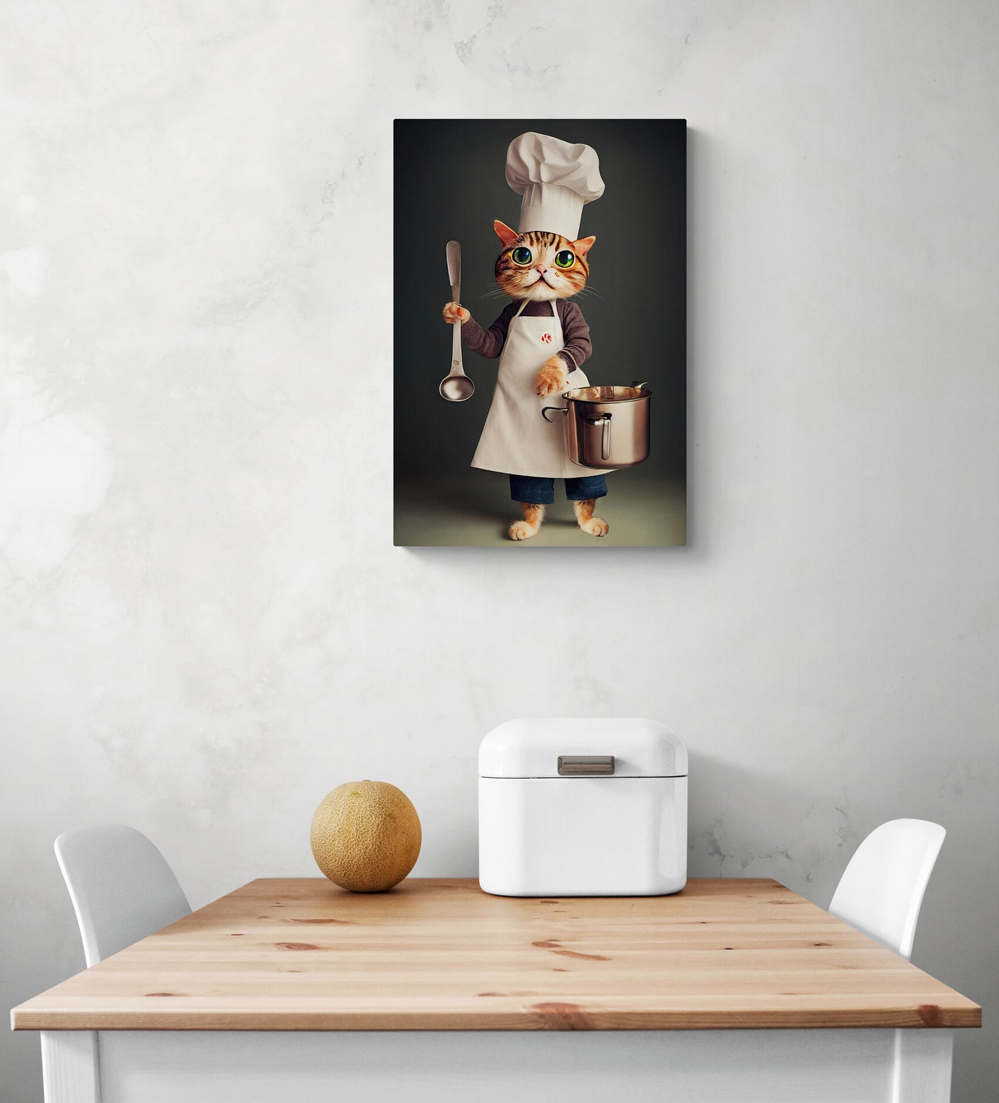 Un tableau cuisine original représentant un gros chat en tenue de chef tenant des ustensiles pour préparer un repas est sur le mur blanc d'une cuisine. Une louche et une casserole sont dans la main du chef. Une table à manger en bois et deux chaises se trouvent en dessous. Une corbeille à pain en métal et un melon sont posés dessus